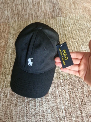 Svart keps från Polo Ralph Lauren - Snygg svart keps från Polo Ralph Lauren med klassisk vit broderad logga framtill och 'POLO' broderat bak. Justerbart spänne baktill för perfekt passform. Tillverkad i bomull för en skön och stilren look.