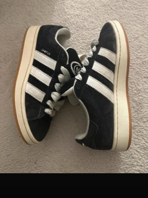 Adidas Campus svarta sneakers - Klassiska Adidas Campus sneakers i svart mocka med vita läderdetaljer och de ikoniska tre ränderna på sidorna. Skorna har vit sula med gummiyta undertill och breda vita skosnören. Adidas-logga på plösen och sidan.