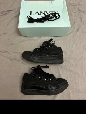 Svarta Lanvin sneakers med box - Säljer ett par helsvarta Lanvin sneakers med chunky sula och breda snören. Skorna har detaljer i både mocka och läder samt metallöljetter. Kommer med originalkartong och extra skosnören. Perfekta för dig som gillar en clean och stilren look.