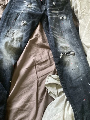Svarta distressed jeans Dsquared2 - (HELT NYA) Säljer ett par svarta jeans från Dsquared2 med slitningar, hål och färgstänk för en riktigt edgy look. Jeansen har en smal passform och coola detaljer som ger dem en unik streetwear-vibe. Perfekta för dig som gillar statement-plagg.