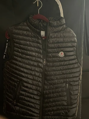 Svart dunväst från Moncler - Snygg svart dunväst från Moncler med glansig finish och klassisk Moncler-logga på bröstet. Västen har två dragkedjefickor framtill och hög krage. Perfekt för lager-på-lager och streetstyle.          Använt 1-2 gånger bara äkthetsbevis finns 