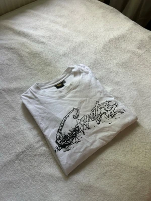 Vit t-shirt från Corteiz - Säljer en vit t-shirt från Corteiz med ett stort svart graffiti-inspirerat tryck på framsidan. Klassisk rund hals och korta ärmar. Perfekt för dig som gillar streetwear och vill sticka ut med en unik design.