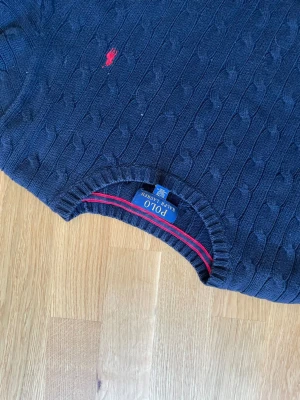 Mörkblå kabelstickad tröja Polo Ralph Lauren - Kabelstickad mörkblå tröja från Polo Ralph Lauren med klassisk röd logga broderad på bröstet. Tröjan har rund halsringning, ribbade muddar och är tillverkad i mjukt material. Perfekt för dig som gillar stilren och tidlös design.