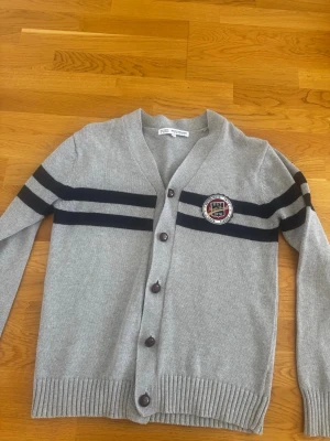 Grå kofta med svarta ränder - Stilren grå kofta från northbrook med svarta dubbla ränder över bröstet och armarna. Koftan har V-ringning, knäppning framtill med mörka knappar och en broderad patch på bröstet. Ribbstickade muddar vid ärmslut och nederkant. Perfekt för en sportig och avslappnad look.