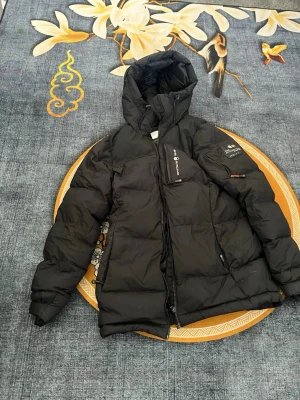 Svart dunjacka från Sail racing - Snygg svart dunjacka från Sail racing med huva och dragkedja framtill. Jackan har flera fickor, patch på ärmen och diskreta logodetaljer. Perfekt för kalla vinterdagar och riktigt varm tack vare dunfyllningen. Köpt från hemsidan runt 3500kr, använd ett par gånger. Pris kan diskuteras. Passar 13-16 år.