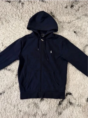 Mörkblå hoodie från Polo Ralph Lauren - Mörkblå hoodie med dragkedja från Polo Ralph Lauren. Klassisk design med huva och dragsko, två fickor framtill och broderad logga på bröstet. Tillverkad i mjukt bomullsmaterial som är skönt att ha på sig.