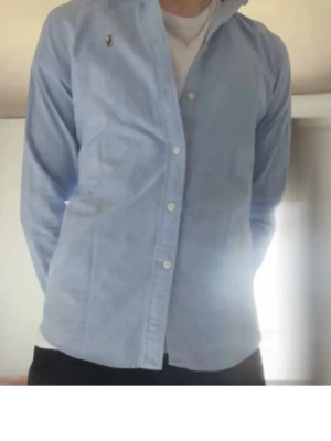 Ljusblå skjorta från Ralph Lauren - Säljer en stilren ljusblå skjorta från Ralph Lauren i slim fit-modell. Skjortan har klassisk krage, knappar framtill och en liten broderad logga på bröstet. Perfekt för dig som gillar en clean och tidlös look. Obs det står 6 på storleken som motsvarar s.