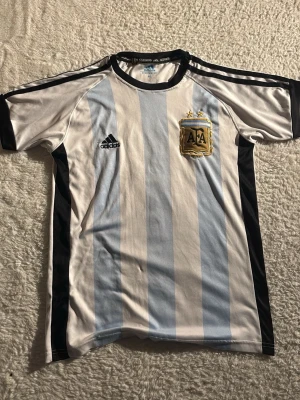 Argentina Adidas fotbollströja - Säljer en klassisk Argentina fotbollströja från Adidas med ljusblå och vita vertikala ränder, svart Adidas-logga och AFA-emblem med två stjärnor på bröstet. Tröjan har svarta detaljer på axlar och sidor samt rund hals. Perfekt för dig som älskar fotboll och vill stötta landslaget.