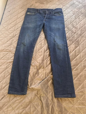 Blå raka jeans från Diesel - Säljer ett par klassiska blå jeans från Diesel med raka ben och snygga slitningar på låren. Jeansen har fem fickor och detaljer i metall, samt Diesel Industry-märke på myntfickan. Perfekta för en avslappnad och trendig stil.