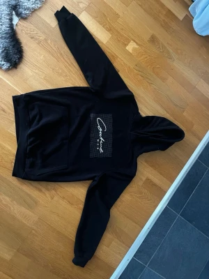 Svart hoodie från new Yorker - Snygg svart hoodie från new Yorker med stor magficka och vit logga på bröstet. Klassisk huva och ribbade muddar vid ärmslut och nederkant. Perfekt för en avslappnad och trendig stil.