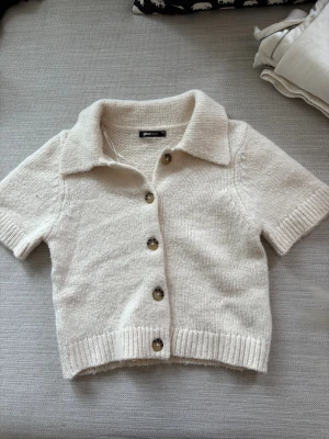 Beige stickad kortärmad kofta Gina Tricot - Kortärmad stickad kofta i beige från Gina Tricot. Koftan har krage, ribbade muddar och knappar framtill i brunmelerad plast. Perfekt till lager på lager och enkel att styla med olika outfits. Tröjan har endast använts en gång.  
