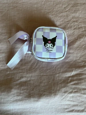 Kuromi - Söt liten väska med Kuromi-motiv från Sanrio. Väskan har ett lila och vitt rutigt mönster, en stor rosett i satin på dragkedjan och är fyrkantig i formen. Perfekt för dig som gillar kawaii-stil och vill ha något must have i den
