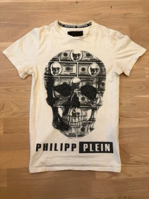 Vit Philipp Plein t-shirt  - Cool vit t-shirt från Philipp Plein med ett stort svart tryck av en döskalle och dollarsedlar på framsidan. T-shirten har rund hals, korta ärmar och logotypen tryckt nedtill. Storlek S!