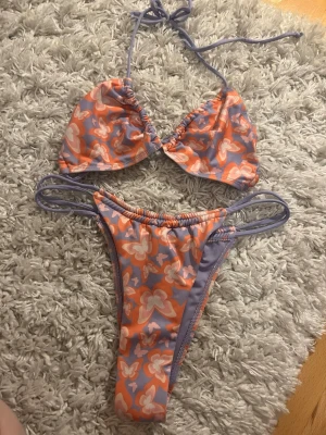 Bershka fjäril Lila och orange bikini - Trendig bikini med trekantsöverdel och smala axelband. Snyggt blommigt mönster i lila, orange och vitt. Underdelen har dubbla sidoband för extra detalj. Perfekt för sommaren och strandhäng!