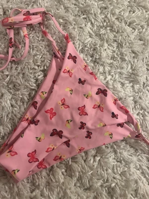 Rosa bikini med fjärilsmönster - Supersöt rosa bikini med små fjärilar i olika färger. Klassisk trekantsmodell med knyt i sidan och runt nacken. Perfekt för sommaren och strandhäng. Materialet känns mjukt och följsamt, troligtvis syntet. Fräsch och lekfull design!