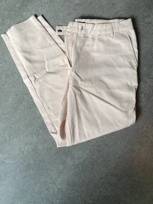 Ljusbeige chinos med raka ben - Snygga ljusbeige chinos med raka ben och klassiska fickor fram samt en passpoalficka bak. Byxorna har bälteshällor och stängs med dragkedja och knapp. Tillverkade i ett mjukt och lätt bomullsmaterial som ger en clean och fräsch look. En osynlig liten dragkedja vid ankeln.
