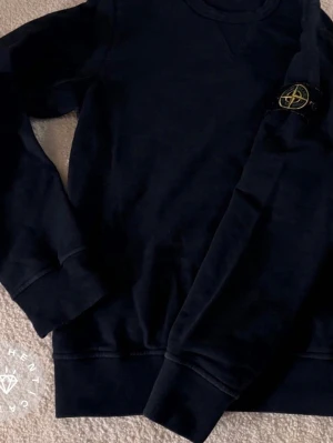  sweatshirt från Stone Island - Säljer en mörkblå sweatshirt från Stone Island med klassisk rund halsringning och ribbade muddar. Tröjan har den ikoniska Stone Island-loggan på ärmen och är tillverkad i mjukt bomullsmaterial. Perfekt för en clean och stilren look.