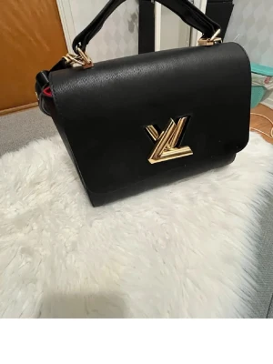 Svart Louis Vuitton handväska i läder - Snygg svart handväska från Louis Vuitton i strukturerat läder med guldfärgade metalldetaljer och ikoniskt LV-spänne framtill. Väskan har både ett topphandtag och en justerbar axelrem för olika stilar. Perfekt accessoar för dig som gillar lyxig design. Har dock haft den i en bra tag så lådan och kvitton till väskan saknas. Men pris kan diskuteras💕