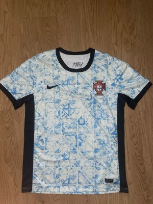 Portugal Nike fotbollströja blåvit - Säljer en Portugal fotbollströja från Nike i vitt med blått mönster och svarta detaljer. Tröjan har rund hals, korta ärmar och Portugal-emblemet på bröstet. Perfekt för dig som vill stötta landslaget med stil.