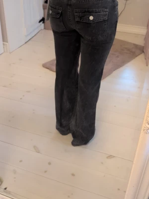 Svarta bootcut jeans med fickor - Säljer ett par svarta bootcut jeans med utsvängda ben och stora bakfickor med lock och knappdetaljer. Jeansen har hög midja och klassisk femficksdesign. Perfekta för en trendig och avslappnad stil.