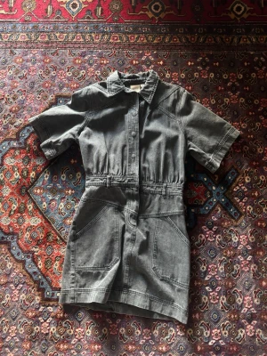 Grå denim klänning med kort ärm - Snygg grå klänning  i denim med korta ärmar, markerad midja och stora fickor framtill. Klassisk skjortkrage och dragkedja framtill. Perfekt för dig som gillar en avslappnad men trendig look. 