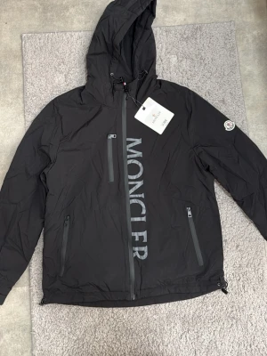 Svart Moncler vindjacka med huva - Snygg svart vindjacka från Moncler med stor logga på bröstet och patch på ärmen. Jackan har huva, tre dragkedjeförsedda fickor och justerbar nederkant. Tillverkad i lätt och slitstarkt material, perfekt för blåsiga dagar.