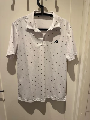 Vit pikétröja från Adidas - Snygg vit pikétröja från Adidas med små svarta mönster över hela tröjan och klassisk krage. Tröjan har korta ärmar och är tillverkad i ett lätt och funktionellt material som passar perfekt till sport eller träning. Golfpike men funkar som vanlig. Helt ny.