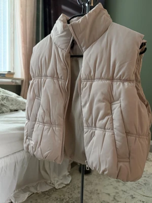Beige puffad och croppad dunväst med hög krage - Snygg beige dunväst med puffad design och hög krage. Västen har två sidofickor och dragkedja framtill. Perfekt för lager på lager. Materialet är quiltat. Använd ett fåtal gånger. 