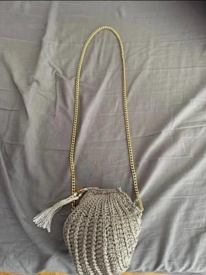 Beige virkad handväska med kedja - Säljer en beige virkad handväska med guldig kedjerem och dekorativ tofs. Väskan har en rundad form och är gjord i grovt garn som ger en trendig och unik look. Perfekt accessoar för dig som gillar boho-stil och vill sticka ut. Köpt i Egypten 