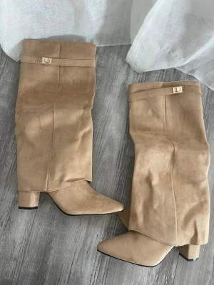 Beige knähöga boots med klack - Snygga beige stövlar i mockaimitation med bred klack och spetsig tå. Stövlarna har dekorativt spänne upptill och en slouchy passform som ger en avslappnad men trendig look. Perfekta att styla till jeans eller kjol för en cool outfit. Aldrig använt! Mer som en 39/40 än 41