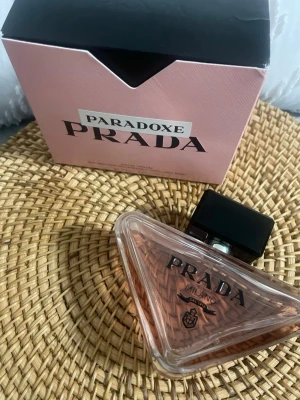 Prada Paradoxe Eau de Parfum - Prada Paradoxe Eau de Parfum med en unik trekantig glasflaska och svart fyrkantigt lock. Doften har en elegant rosa ton och kommer i en stilren rosa kartong med svart text. Perfekt för dig som gillar exklusiva och moderna parfymer.
