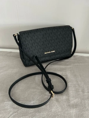 Svart axelväska Michael Kors - Snygg svart axelväska från Michael Kors med justerbar axelrem och guldfärgade detaljer. Väskan har ett klassiskt MK-mönster över hela ytan och stängs med magnetlås. Perfekt storlek för det viktigaste och stilren design som passar till många outfits.