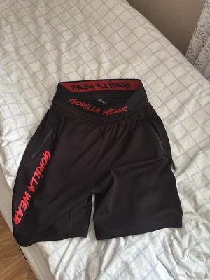 Svarta shorts från Gorilla Wear - Säljer ett par svarta träningsshorts från Gorilla Wear med röda detaljer och logga på både midjan och benet. Shortsen har dragkedjeförsedda fickor och elastisk midja för skön passform. Perfekta för gymmet eller annan träning.