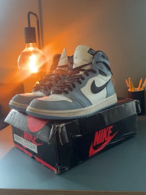 Nike Air Jordan 1 High OG blå/vit - Säljer ett par Nike Air Jordan 1 High OG sneakers i blått och vitt med svarta detaljer. Klassisk hög siluett med svart swoosh och vita paneler. Skorna har svart snörning och blå yttersula. Perfekta för dig som gillar streetwear och ikoniska modeller.