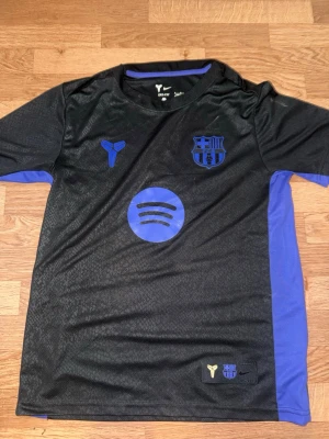 Svart Barcelona Nike matchtröja - Säljer en svart Barcelona matchtröja från Nike med blå detaljer och loggor. Tröjan har klubbmärke, sponsorlogga (Spotify) och Nike-logga på bröstet. Tillverkad i ett lätt och ventilerande material, perfekt för fotboll eller träning.