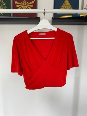 Röd croppad topp med v-ringning - Snygg röd croppad topp med djup v-ringning och korta, lösa ärmar. Toppen har ett draperat fall och markerad midja som framhäver kurvor!