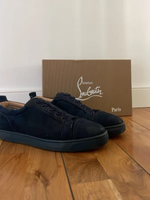 Svarta sneakers från Christian Louboutin - Stilrena svarta sneakers från Christian Louboutin i mocka med svarta snören och diskret design. Låg modell med klassisk siluett och svart yttersula. Perfekta för dig som gillar exklusiva och minimalistiska skor. Lite slitna som du ser på bilderna!