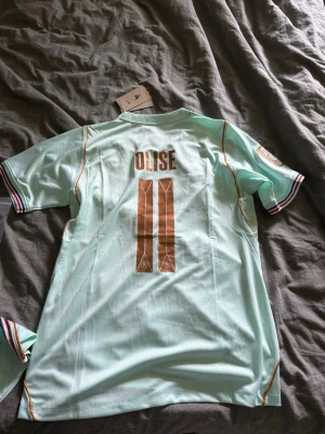 Mintgrön fotbollströja Olise nr 11 - Mintgrön fotbollströja med namnet Olise och nummer 11 i kopparfärg på ryggen. Tröjan har korta ärmar med randiga muddar i vitt, rött och svart samt tunna orange detaljer längs sidorna. Perfekt för dig som gillar sportig stil. Bild på framsidan finns på andra annonsen med shortsen. Skick 10/10 och är aldrig använd. Hör av er vid frågor! 