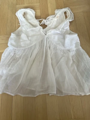 Vit linnetopp med knyt i ryggen - Säljer en luftig och söt vit linnetopp med tunna axelband och knyt i ryggen. Toppen har en flowy passform och är tillverkad i ett skirt, lätt material som passar perfekt till varma dagar. Enkel och stilren design som är lätt att matcha.