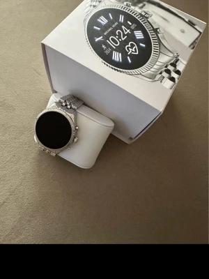 Michael Kors silverfärgad smartklocka - Snygg smartklocka från Michael Kors i silverfärgat rostfritt stål. Klockan har en rund urtavla med digital display och klassisk länkarmband. Stilig design med räfflad kant runt skärmen och flera knappar på sidan för enkel navigering.