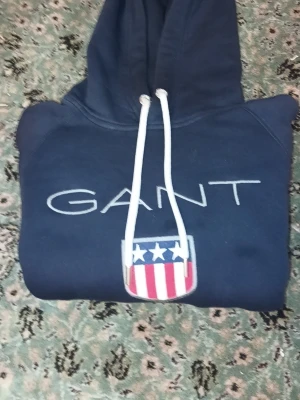 Mörkblå hoodie från GANT - Snygg mörkblå hoodie från GANT med vit logga och amerikanskinspirerat sköldmärke på bröstet. Hoodien har vit dragsko i huvan och en klassisk magficka. Perfekt för dig som gillar stilrena och sportiga plagg. Anvönt ENFAST 1 GÅNG!! FKCK Den som födsdegasprest. Storlrk L men passar M och S men mest M