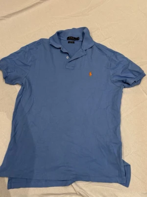 Blå pikétröja från Polo Ralph Lauren - Klassisk blå pikétröja från Polo Ralph Lauren med den ikoniska broderade loggan i orange på bröstet. Tröjan har krage, korta ärmar och knappar vid halsen. Perfekt för en avslappnad men ändå stilren look.