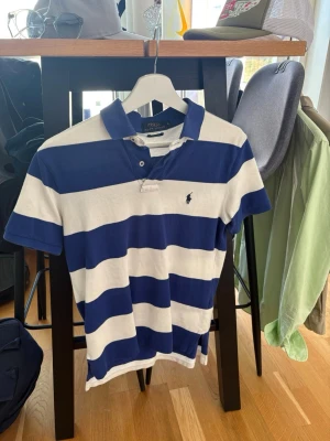 Randig pikétröja från Polo Ralph Lauren - Klassisk pikétröja med breda blå och vita ränder från Polo Ralph Lauren. Tröjan har korta ärmar, krage och knappar vid halsen samt den ikoniska broderade loggan på bröstet. Perfekt för en avslappnad och stilren look.