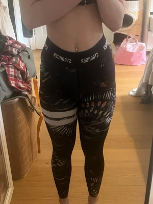 Svarta Stronger leggings med mönster - Säljer ett par svarta leggings från Stronger med coolt abstrakt mönster i vitt, orange och grått. Hög midja med elastisk linning där det står 'STRONGER' runtom. Tight passform och stretchigt material som sitter snyggt på kroppen.