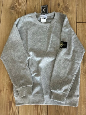Grå sweatshirt från Stone Island - Säljer en stilren grå sweatshirt från Stone Island med klassisk rund halsringning och ribbade muddar. Tröjan har den ikoniska Stone Island-loggan på vänster ärm och är tillverkad i mjuk bomull. Perfekt för en avslappnad och trendig look.