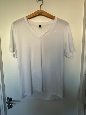 Vit v-ringad t-shirt från G-Star RAW XL - Säljer en stilren vit t-shirt från G-Star RAW med v-ringning och diskret logga på bröstet. T-shirten är kortärmad och gjord i mjuk bomull för en skön känsla. Perfekt till jeans eller shorts för en clean look. En liten fläck vid lappen på ryggen