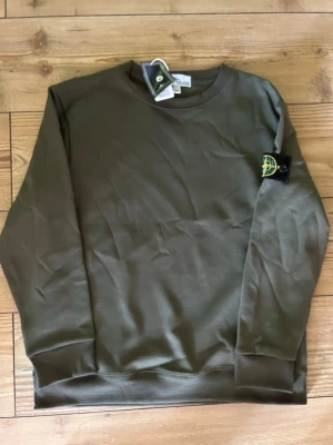 Olivgrön sweatshirt från Stone Island - Snygg olivgrön sweatshirt från Stone Island med klassisk rund hals och ribbade muddar. Tröjan har den ikoniska Stone Island-loggan på vänster ärm och är tillverkad i mjuk bomull. Perfekt för en clean och avslappnad stil.