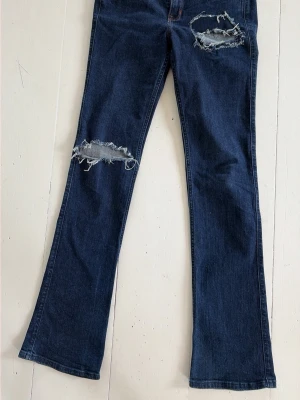 Mörkblå bootcut jeans med slitningar - W27, L33. Snygga mörkblå bootcut jeans med stora slitningar på låret och knät. Jeansen har klassisk femficksmodell och är tillverkade i denim med lätt stretch. Perfekta för en avslappnad och trendig look.