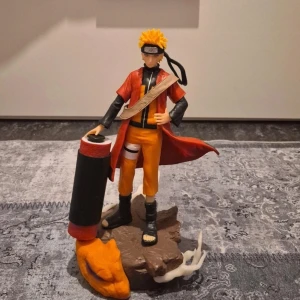 Naruto Uzumaki cosplayfigur - Detaljerad cosplayfigur av Naruto Uzumaki från anime-serien Naruto. Figuren har orange byxor, svart tröja, röd mantel och pannband. Tillbehör som stor rulle och bas med rök och rävhuvud ingår. Perfekt för cosplay eller samling.