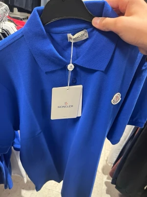 Blå pikétröja från Moncler - Klassisk blå pikétröja från Moncler med krage, tre knappar framtill och broderad logotyp på bröstet. Tillverkad i bomull och har korta ärmar. Perfekt för en stilren och sportig look.
